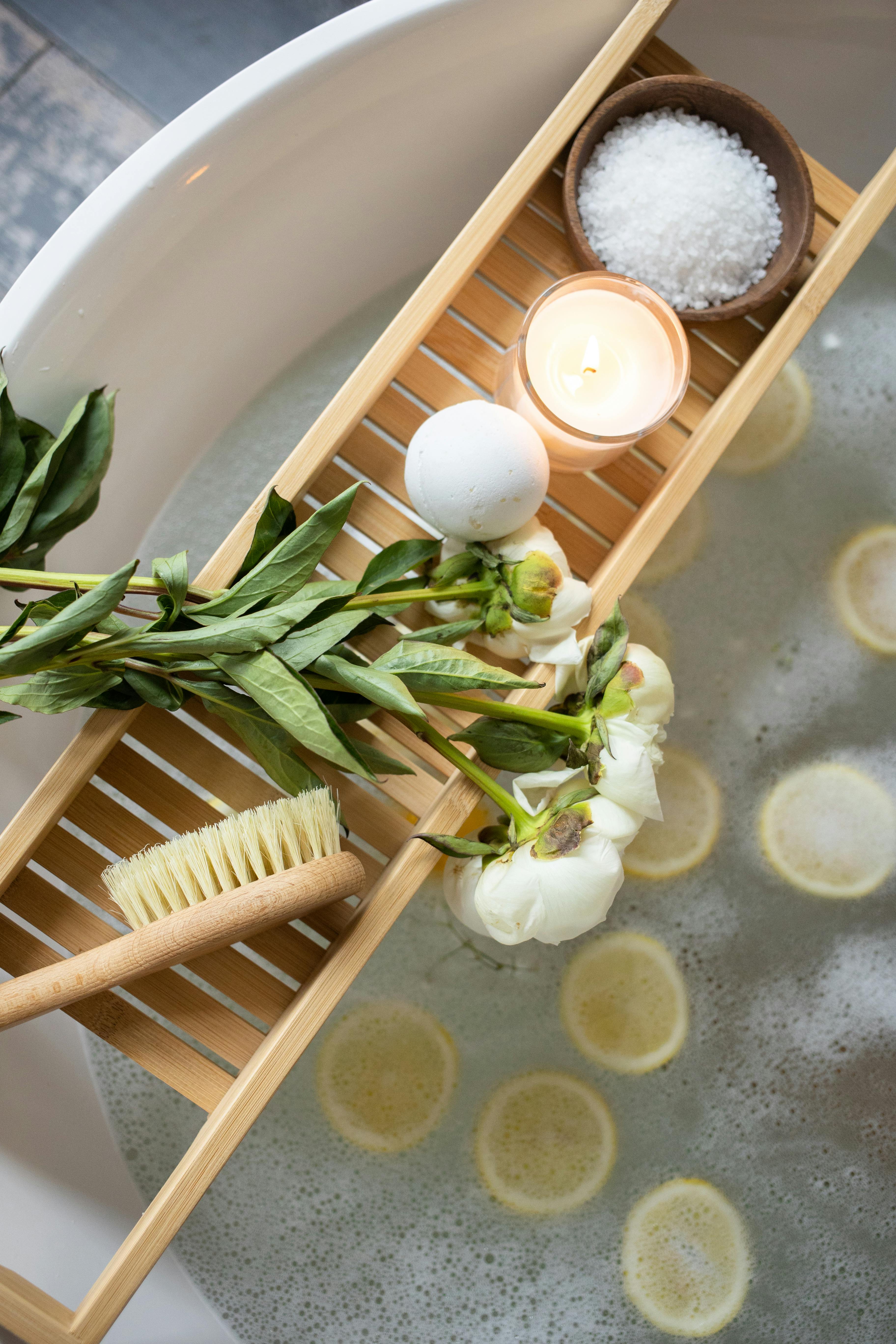 Top view of wooden tray with bath bomb and burning candle placed near salt and flowers on tub with lemon slices on water